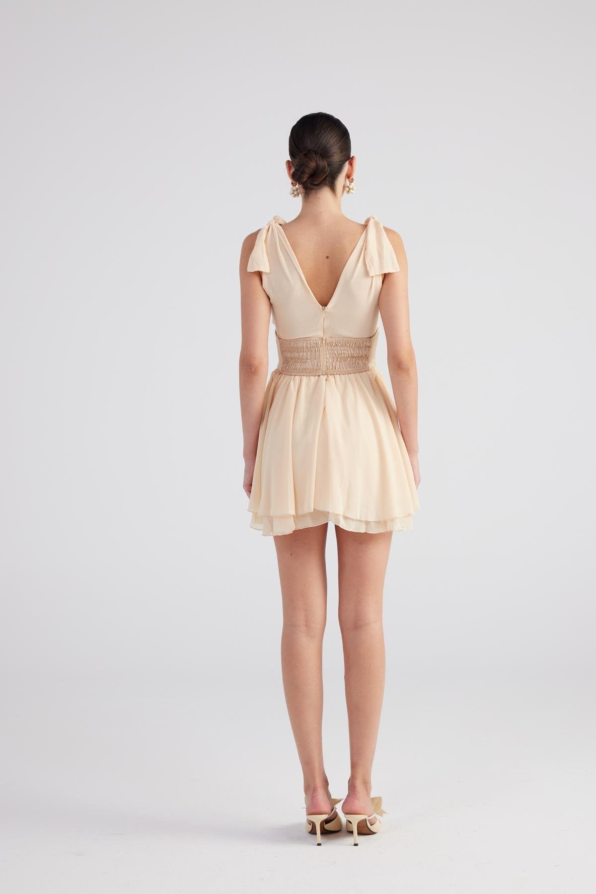 Beige ruched mini dress with V-back detail, shoulder ruffles, and pleated skirt