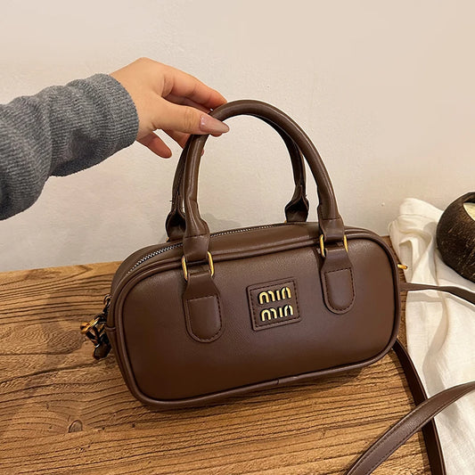 Small brown handbag with top handles, front logo patch, and gold-tone zipper detail